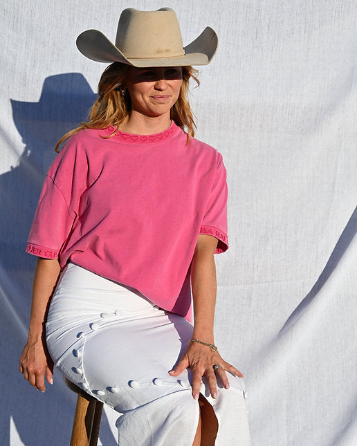 Person wearing a pink shirt and beige cowboy hat sitting on a stool against a white background