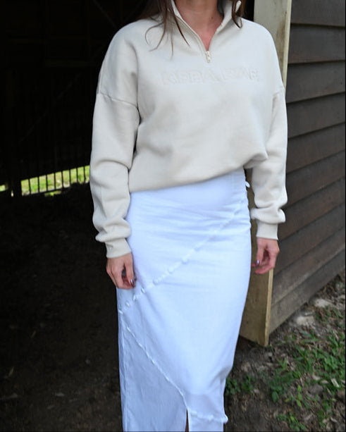 Woman wearing a beige sweater and white skirt standing outdoors.