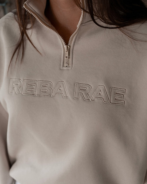 Beige sweatshirt with 'REBA RAE' branding on a person wearing white pants.