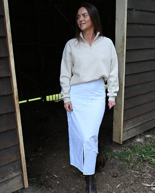 Woman in a beige sweater and white skirt standing in front of a wooden structure.