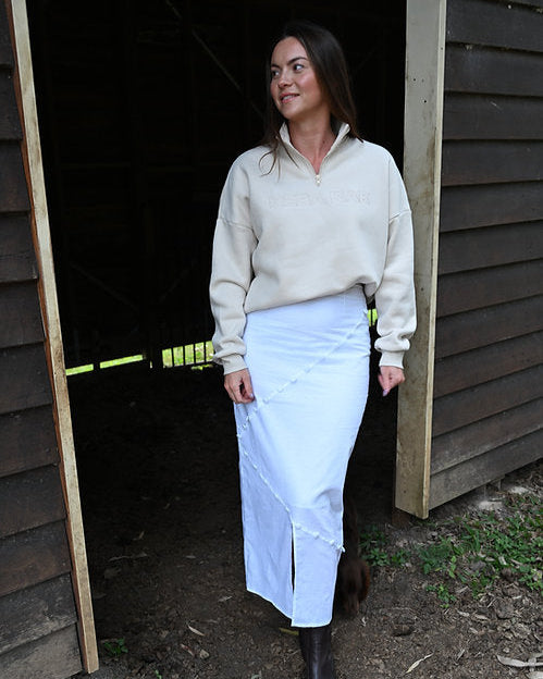 Woman in a beige sweater and white skirt standing in front of a wooden structure.