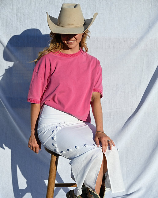 Person wearing a pink shirt, white pants, and cowboy hat sitting on a wooden stool against a white backdrop.