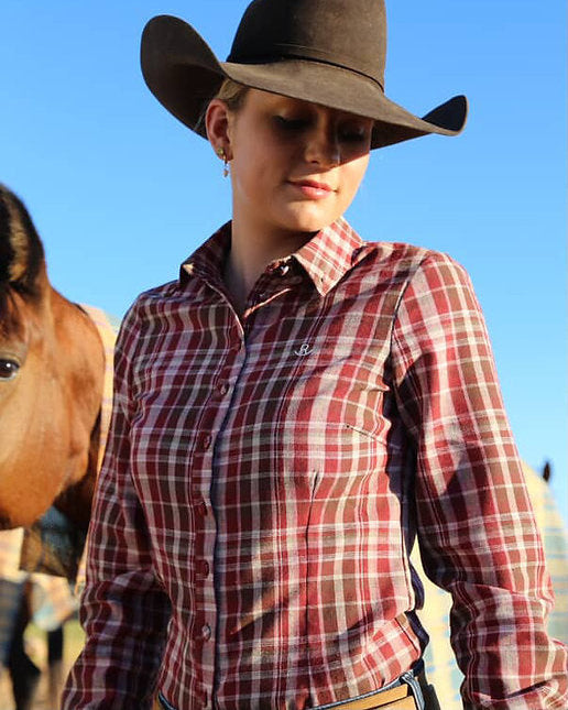 Woman wearing a cowboy hat and plaid shirt standing next to a horse against a blue sky.