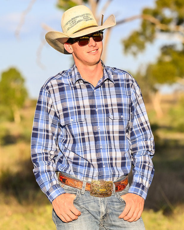 Man wearing a cowboy hat and plaid shirt standing in a field with trees in the background