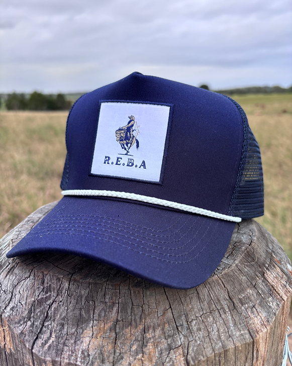 Navy blue cap with a white patch featuring 'R.E.B.A.' logo on a wooden post with a field background.