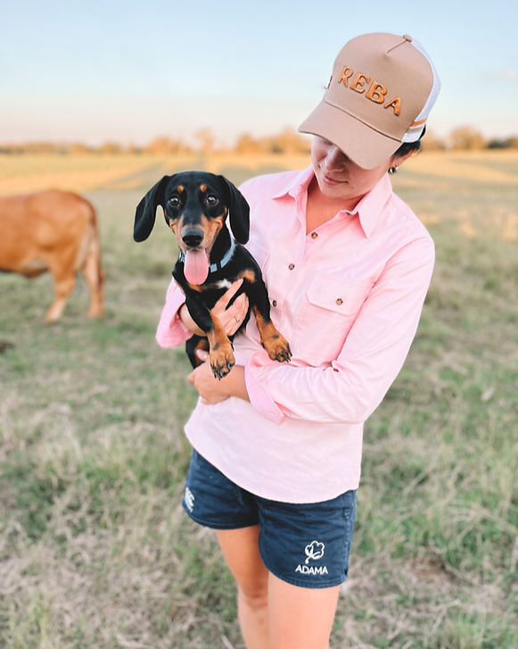 Person holding a small dog in a field wearing a cap and pink shirt.