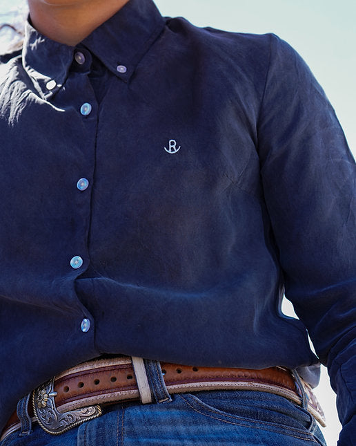 Person wearing a navy blue button-up shirt with a logo, blue jeans, and a brown belt.