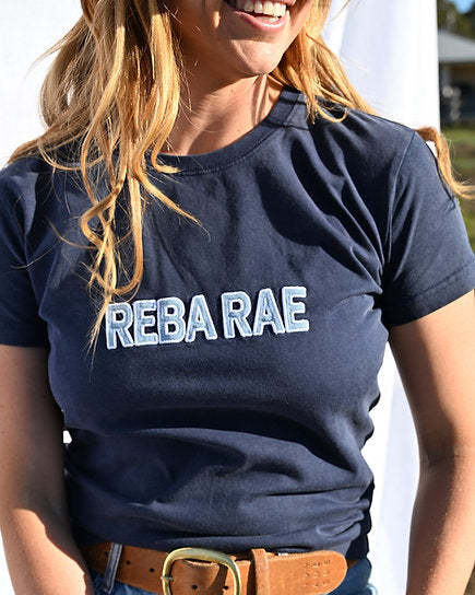 Person wearing a navy blue t-shirt with 'REBA RAE' text, sitting outdoors.