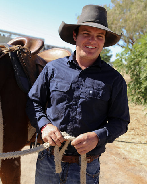 Man in a cowboy hat holding a rope next to a horse, with trees and clear sky in the background.