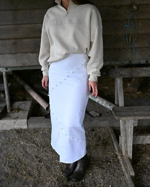 Woman wearing a cream sweatshirt and white apron standing in a rustic setting.