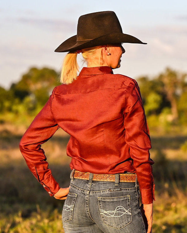 Person wearing a red shirt, black cowboy hat, and jeans standing in a field with trees in the background.