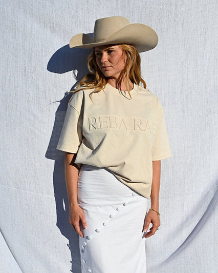 Person wearing a beige 'Rebeka' shirt, white skirt, and cowboy hat against a white background