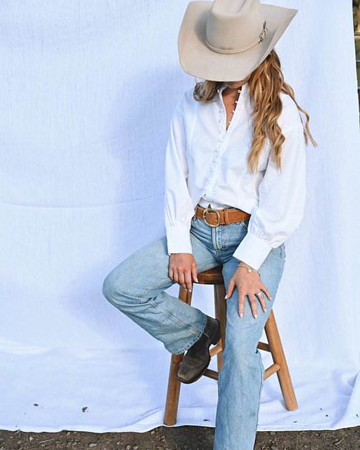 Person wearing a white shirt, blue jeans, and a beige cowboy hat sitting on a wooden stool against a white backdrop.