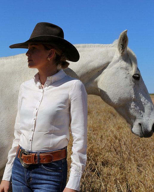 Woman in a white shirt and cowboy hat standing next to a white horse in a field with blue sky.