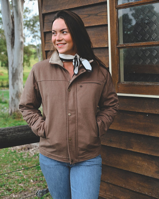 Woman wearing a brown jacket and blue jeans standing in front of a wooden building.