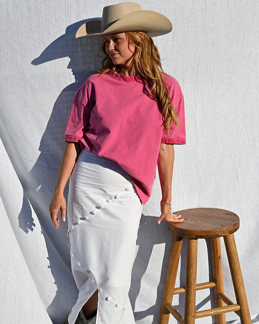 Person wearing a pink shirt and white pants standing next to a wooden stool against a textured white wall.