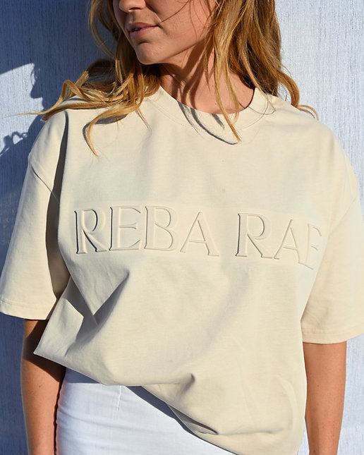 Person wearing a beige t-shirt with 'REBA RAS' text against a blue wall.