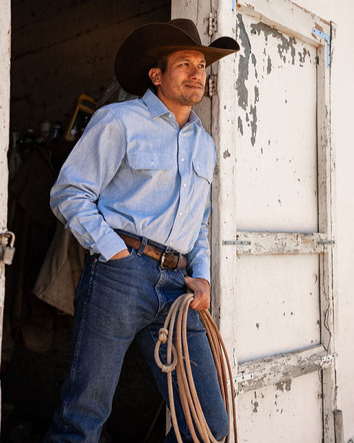 Man in a cowboy hat and light blue shirt holding a rope, standing in a rustic setting.