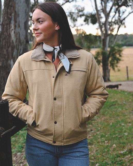 Woman wearing a beige jacket with a scarf, standing outdoors in a natural setting.