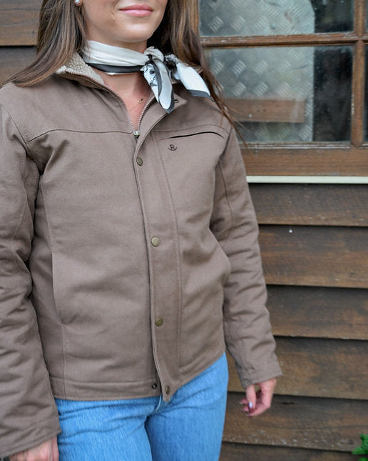 Woman wearing a brown jacket and blue jeans standing in front of a wooden building.