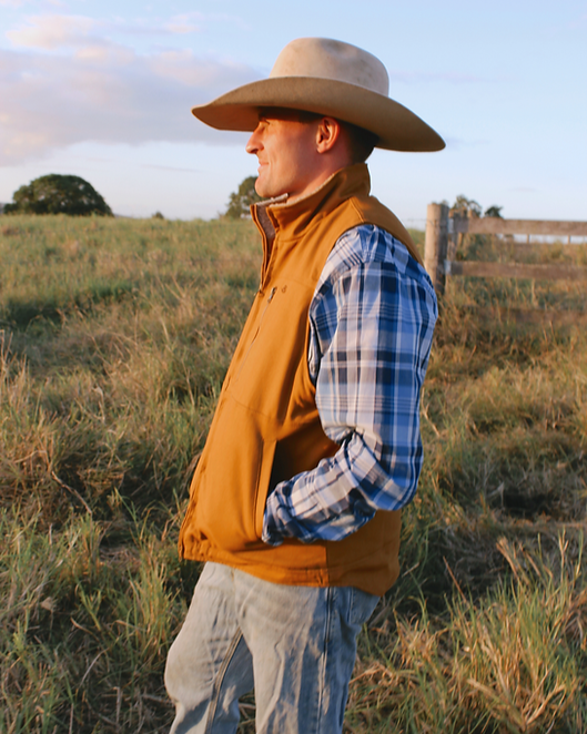 Man wearing a cowboy hat, yellow vest, and plaid shirt standing in a field.