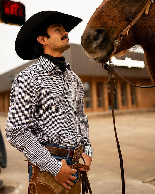 Man in cowboy hat and checkered shirt standing next to a horse in an outdoor setting
