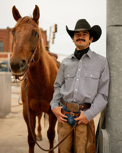 Man in cowboy hat and attire standing next to a horse, leaning against a wall.