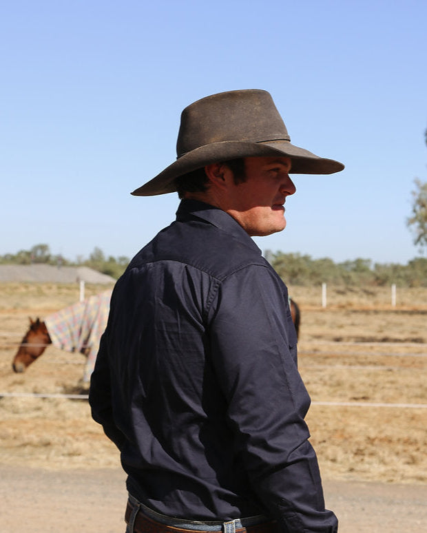 Man wearing a wide-brimmed hat in a rural setting with a horse and trees in the background.