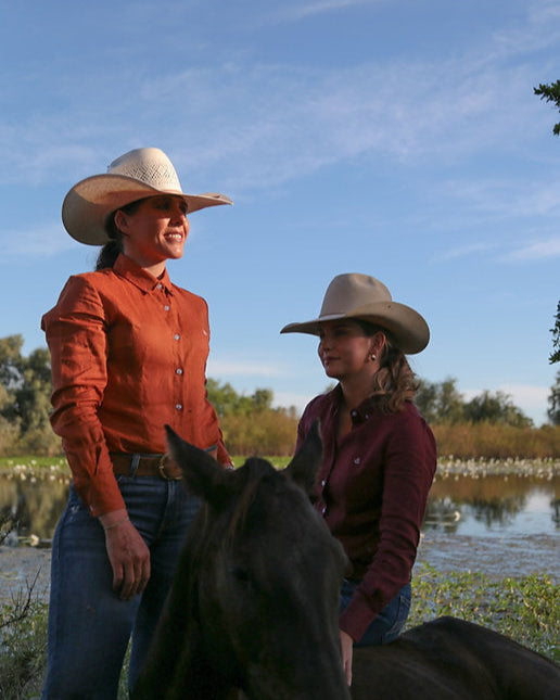 Two women in cowboy hats standing next to horses by a body of water.