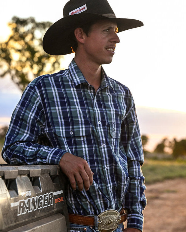 Man in a plaid shirt and cowboy hat standing next to a vehicle with a scenic background